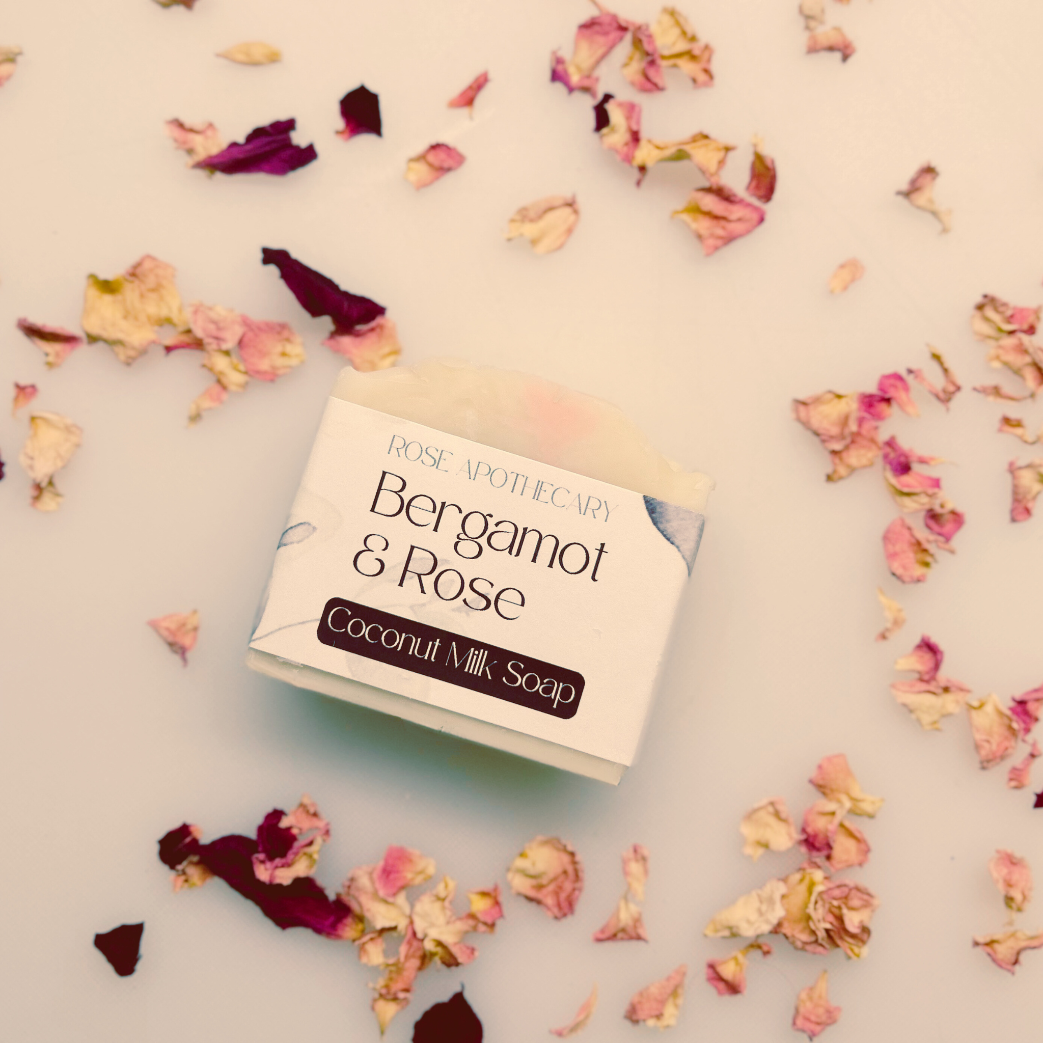 Coconut milk soap bar with 'Bergamot & Rose' branding surrounded by flower petals on a light background