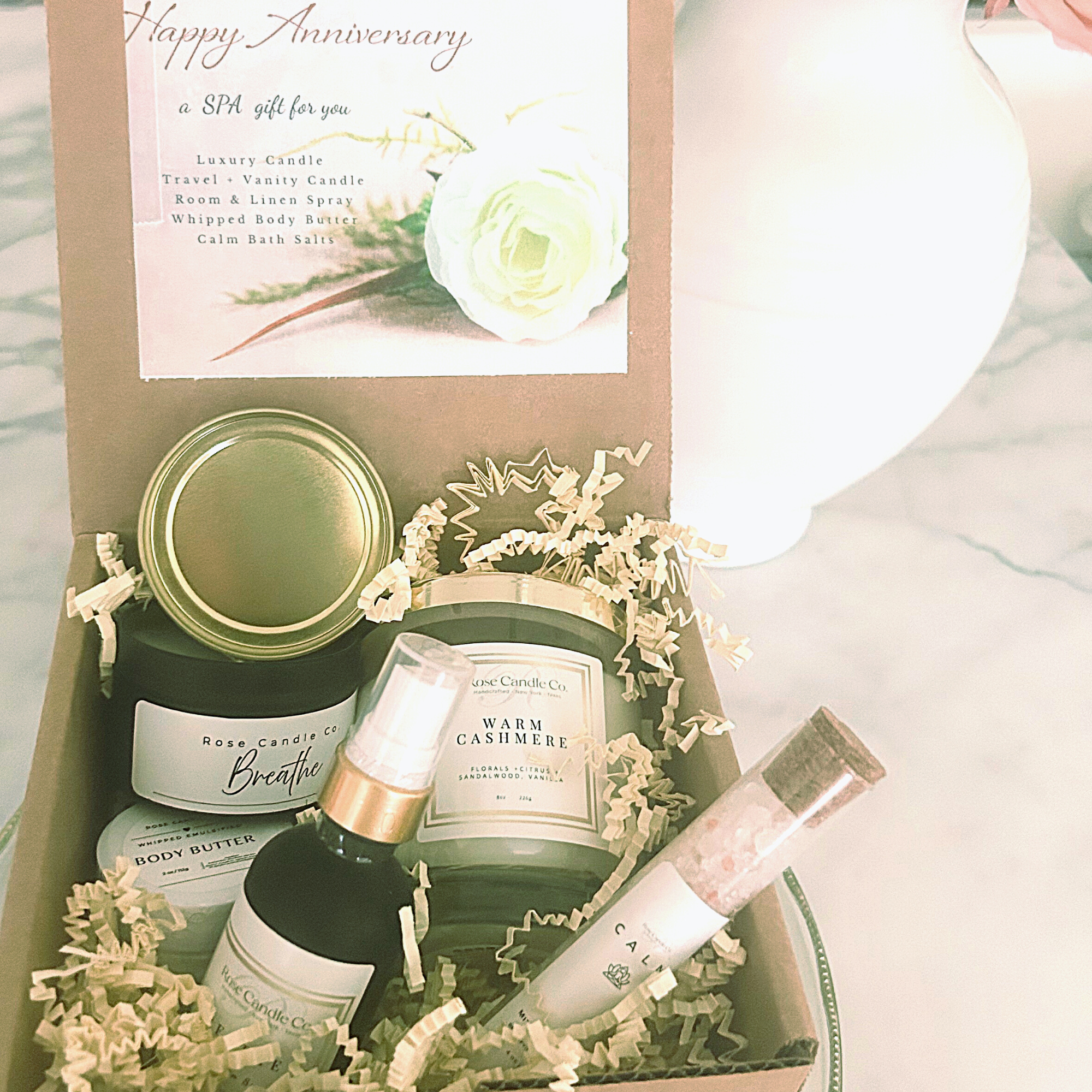 Box filled with confetti surrounding a glass luxury candle, travel candle, body butter , room and linen spray and Dead Sea calming bath salts 